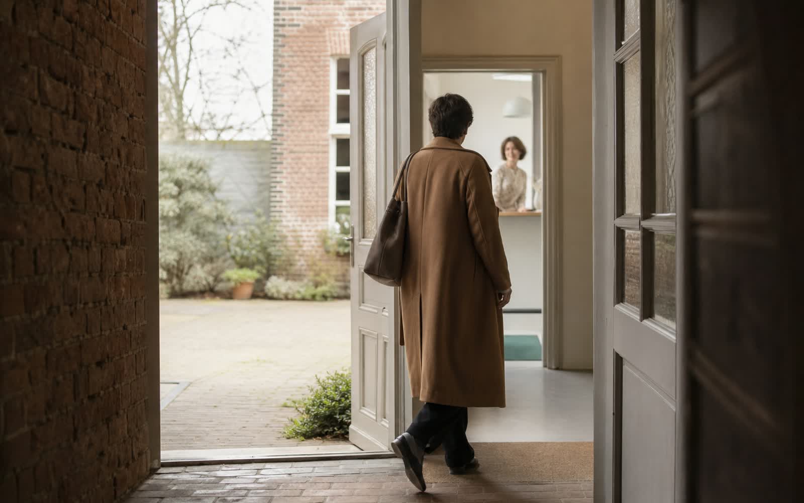 Eine Frau tritt durch eine offene Tür in einen hellen Hof, aus einem dunkleren Flur gesehen
