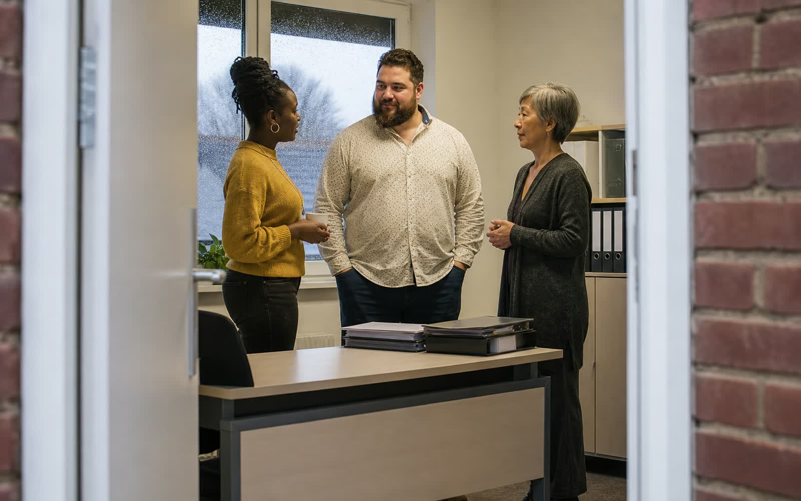 Drei unterschiedlich alte Mitarbeitende sprechen in einem kleinen Büro, gesehen durch eine Tür